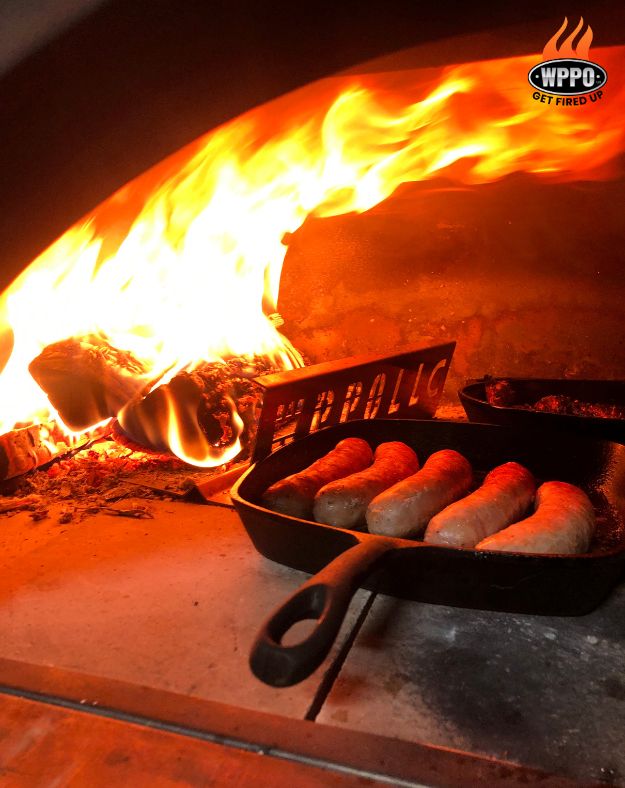 Cast-iron pan cooking inside a WPPO Karma 55-inch commercial wood-fired pizza oven with flames and hot coals