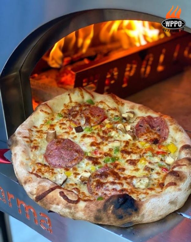 Pizza being removed from a WPPO Karma 32-inch wood-fired pizza oven with flames visible inside