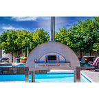 WPPO Karma 55-inch commercial wood-fired pizza oven in a backyard poolside setting with stainless steel chimney