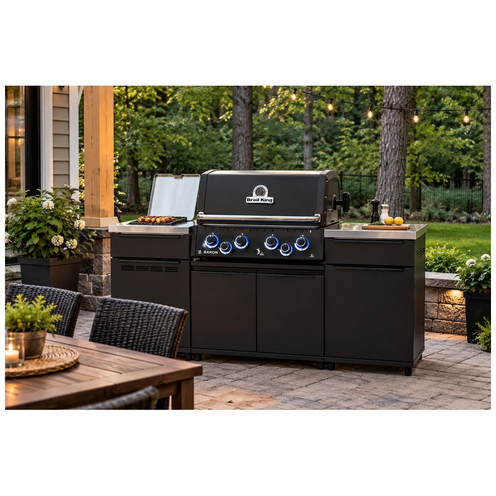 Broil King Baron 490i Pro IR Shadow outdoor kitchen island on a stone patio in a wooded backyard, featuring black cabinetry, stainless steel countertops, an infrared side burner, and illuminated control knobs.