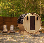 Aleko Liatris cedar barrel sauna with a dark door and shingle roof in a wooded outdoor setting, with two lounge chairs nearby.