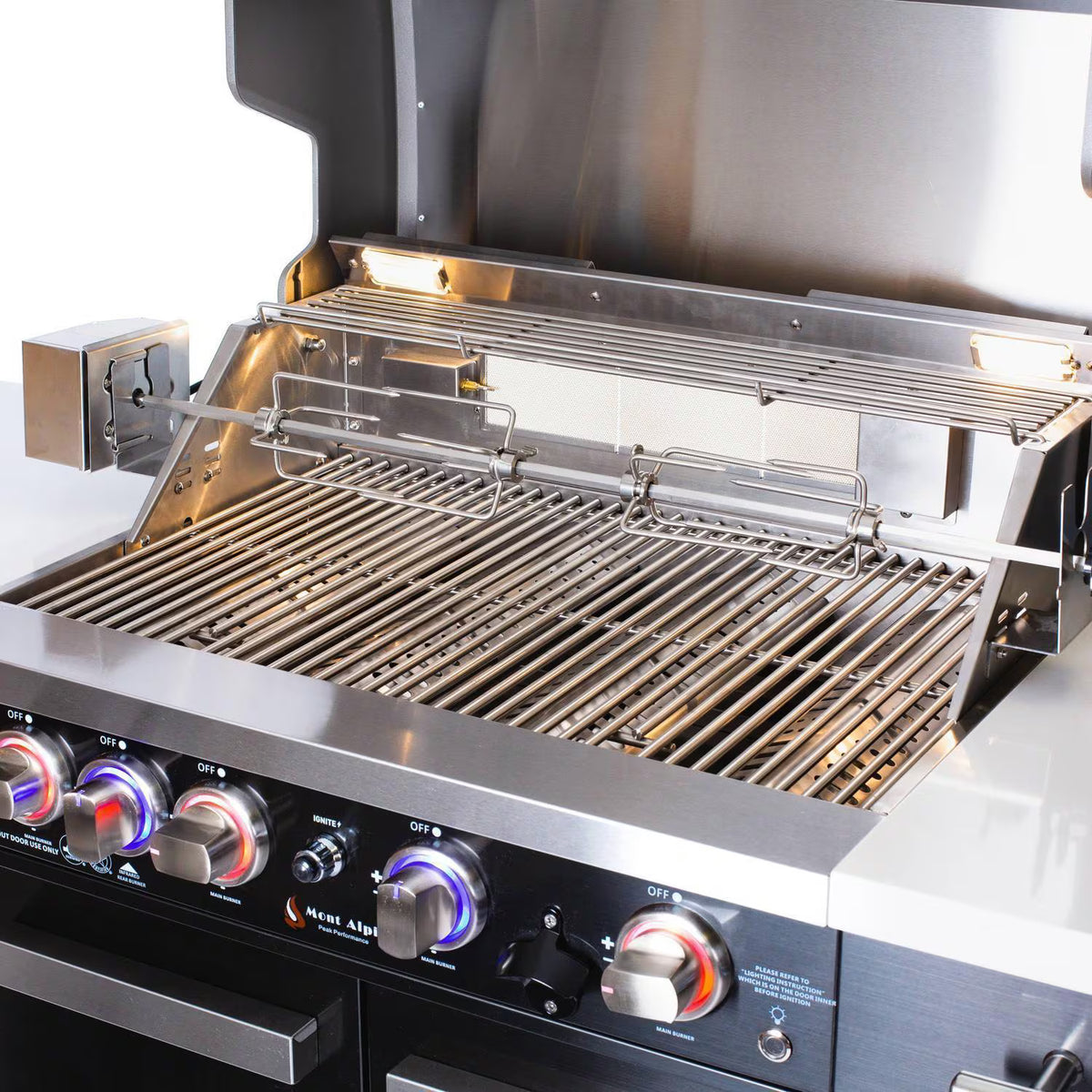 Close-up of Mont Alpi 957 Compact black stainless steel grill with hood open, rotisserie installed, stainless steel cooking grates, and illuminated control knobs